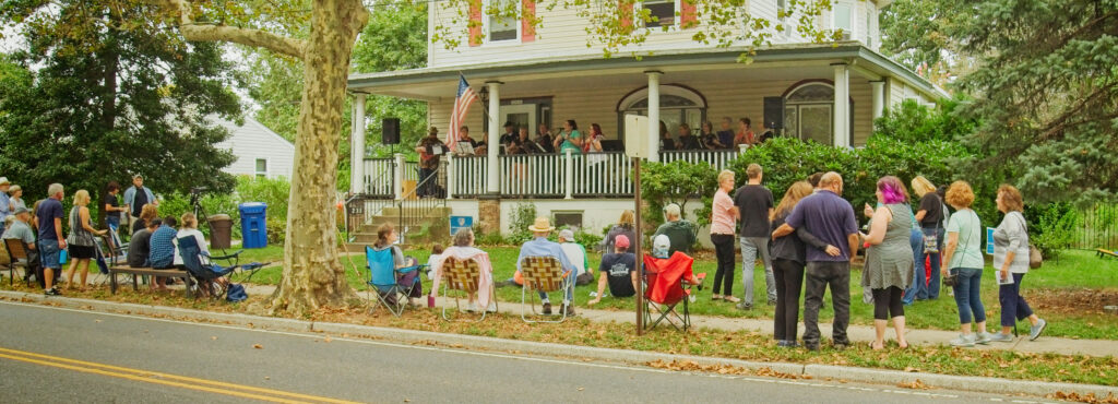 A crowd lines the sidewalk as a band plays on a front porch.