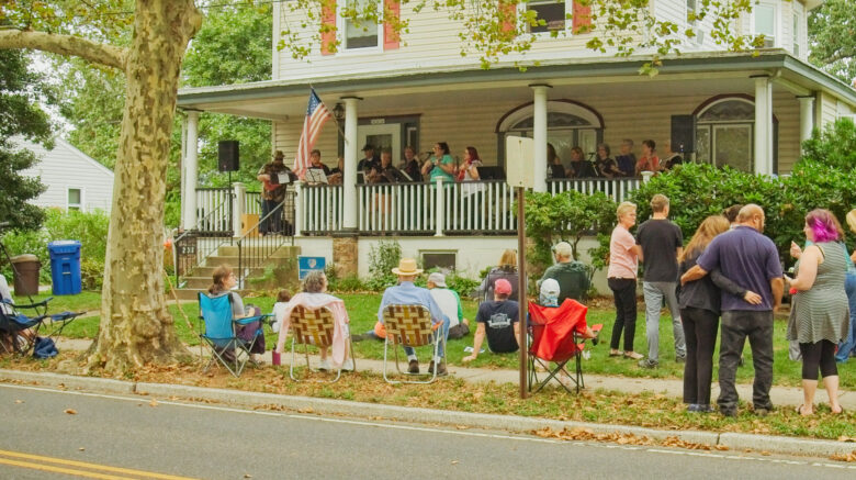 A crowd lines the sidewalk as a band plays on a front porch.
